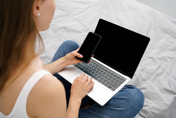Νεαρή γυναίκα κρατώντας τηλέφωνο και laptop με λευκή μαύρη οθόνη, ενώ κάθεται στον καναπέ. Παραμόρφωση φόντου για το διαφημιστικό σας περιεχόμενο. Η έννοια της απομακρυσμένης εργασίας, την εκμάθηση και βλέποντας online μαθήματα και ταινίες. - Φωτογραφία, εικόνα