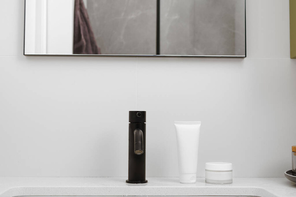 Λευκό mockup ενός σωλήνα κρέμας και ένα γυάλινο βάζο καλλυντικών για θρέψη και ενυδάτωση του δέρματος του προσώπου στο νεροχύτη κάτω από τον καθρέφτη στο μπάνιο. Έννοια των προϊόντων ομορφιάς - Φωτογραφία, εικόνα