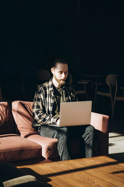 Sakallı bir genç adam bir kafede uzaktan bilgisayarla çalışıyor. Bir bilişim uzmanı sabahları koltukta oturur. Bir kafede internetten okuyan genç bir öğrenci.. - Fotoğraf, Görsel