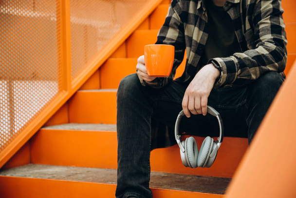 Modern bir ofisin merdivenlerinde oturan bir adam elinde bardak, kulaklık ve dijital saatle. Cihazsız kahve molası. Bilgisayardan uzak dur, dijital detoks. Boşluğu kopyala. - Fotoğraf, Görsel