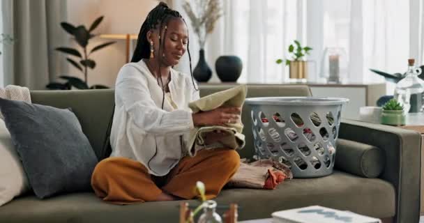 Mosás, pihenés és egy fekete nő hajtogatás mosás egy kanapén a nappaliban az otthonában, hogy rendet rakjon. Mosoly, házimunka és egy boldog fiatal háziasszony takarítja a lakását takarításhoz vagy higiéniához. - Felvétel, videó