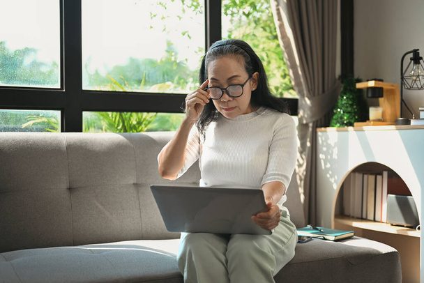 Femme d'âge moyen touchant des lunettes et lisant un e-mail sur un ordinateur portable tout en se relaxant sur le canapé à la maison - Photo, image