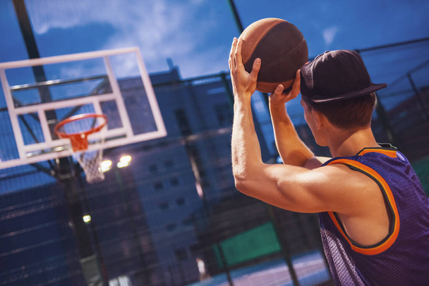 Şık genç basketbolcu şapkalı bir çember ile bir top basketbol sahasına açık havada akşam oynarken ateş ediyor - Fotoğraf, Görsel