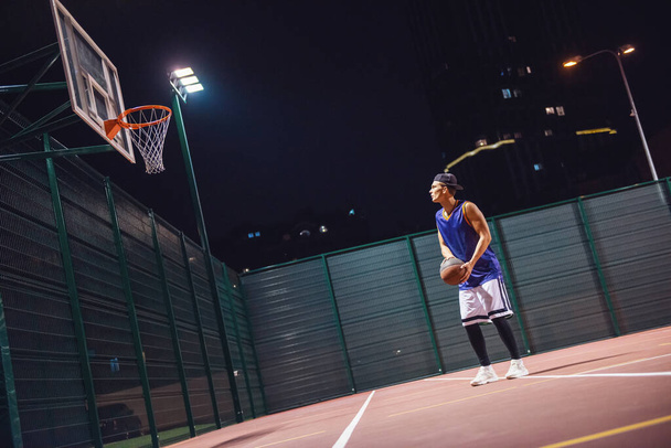 Retrato de larga duración de un joven jugador de baloncesto con estilo en el salto de gorra y disparar una pelota a través del aro mientras juega al aire libre por la noche
 - Foto, imagen
