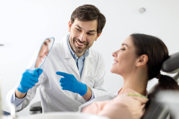 Amistoso dentista masculino profesional que da espejo a su paciente feliz mujer, mostrando el resultado del tratamiento dental o blanqueamiento - Foto, Imagen