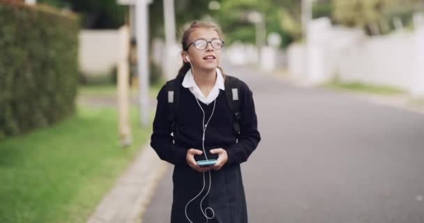 Child, earphones and music in outdoors to school, smartphone and streaming radio or song on commute. Girl, student and walking on road, online chat and ready for education or knowledge at academy. - Footage, Video