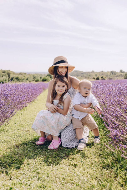 彼女の小さな息子と娘が咲くラベンダー畑を歩いている. ストロー帽子と白いドレスのブルネットの女性. 幸せな母性と子供時代。 ラベンダーフィールドを散策. - 写真・画像