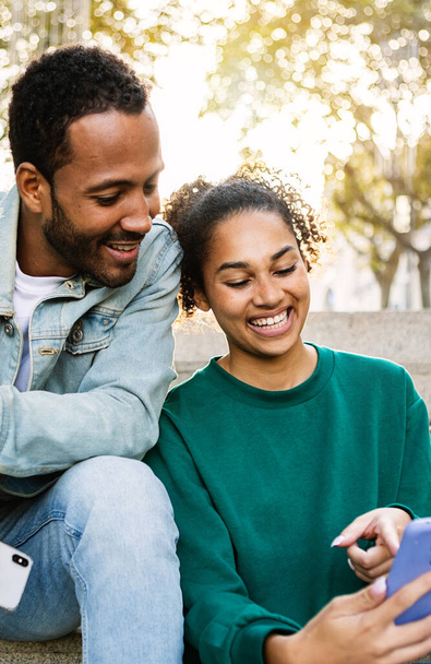Vertikale Aufnahme von zwei jungen Lateinamerikanern, die zusammen im Freien sitzen und ihr Smartphone benutzen. Fröhliche afrikanisch-amerikanische Freunde lachen, während sie auf den Handy-Bildschirm schauen. Social Media-Konzept. - Foto, Bild