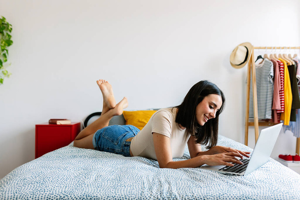 ベッドの上に横たわっているラップトップを使用して若い美しい女性の完全な長さのビュー. 自宅でオンラインで勉強する十代の少女. - 写真・画像
