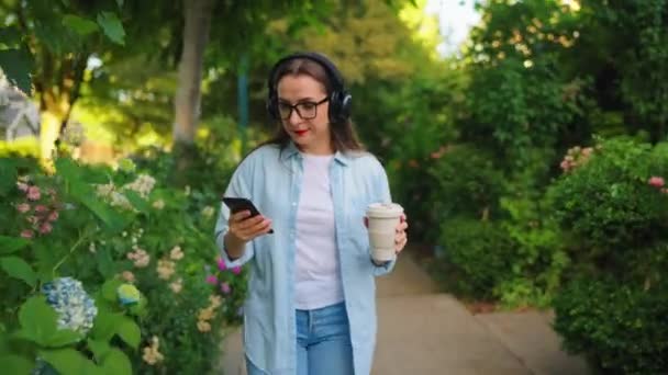 Glückliche Frau mit Brille und Kopfhörer, Kaffee und Smartphone in der Hand, die an Sommertagen durch den blühenden Garten oder die grüne Straße läuft. - Filmmaterial, Video