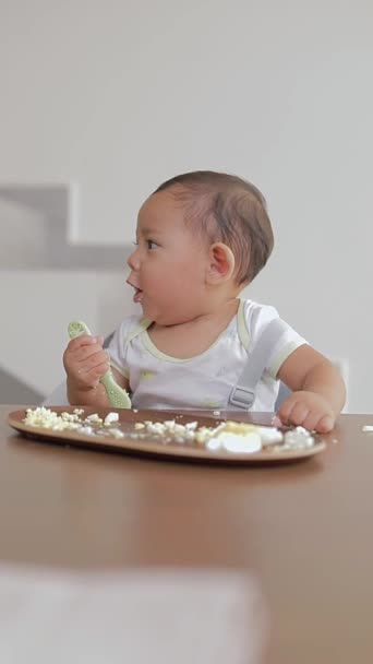 Латиноамериканский ребенок жует ложку во время еды, сидя за столом - Кадры, видео