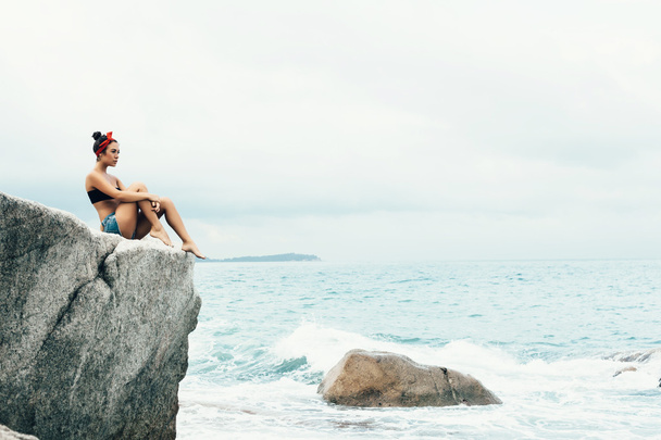 Rear view of  woman sitting and relaxing on a natural coastal rock high up - 写真・画像