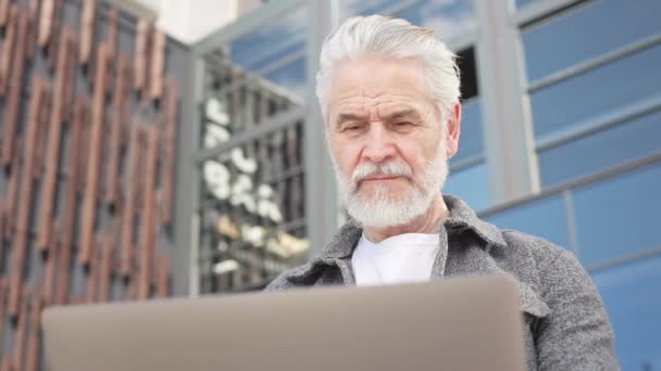 Uomo caucasico barbuto mostrando attenzione mentre seduto su una panchina di legno e tenendo laptop portatile in ambiente esterno. Maturo signore impegnarsi in un lavoro all'aperto con gravi emozioni facciali. - Filmati, video