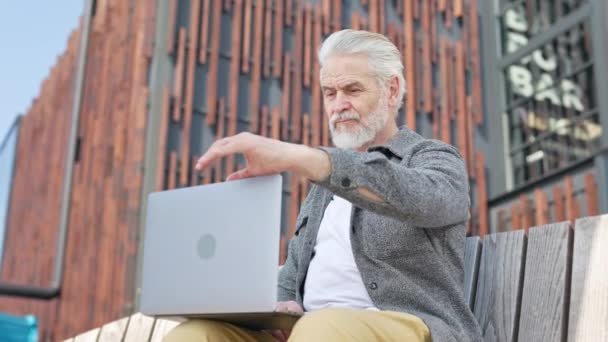 Ein älterer Herr öffnet und begutachtet den modernen Laptop, während er auf einer bequemen Bank im Freien sitzt. Schön gekleideter Mann in Freizeitkleidung bei der Arbeit im Central Park. - Filmmaterial, Video