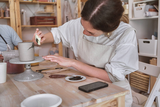 Artiste attentif utilisant une éponge pour peindre une tasse grise alors qu'elle tourne sur une roue de poterie dans un studio ensoleillé - Photo, image