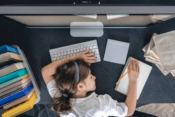 Egy fáradt lány fekszik az asztalon egy számítógép előtt, jegyzetfüzetekkel és könyvekkel, felülnézetből. Egy diák alszik a házi feladata alatt. Távoli tanulás, otthoni tanulás. - Fotó, kép