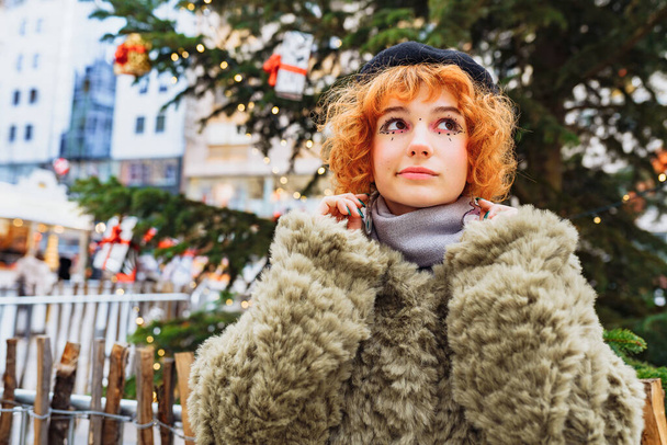 Portrait attractive teenage girl, young woman, with red curly hair, wearing beret, fur coat, with festive makeup against background of Christmas decor in European city - Photo, Image
