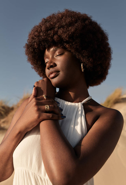 Young sensual black woman with Afro hairstyle in white dress stroking hand tenderly while standing on beach in sunshine with closed eyes - Foto, Imagem