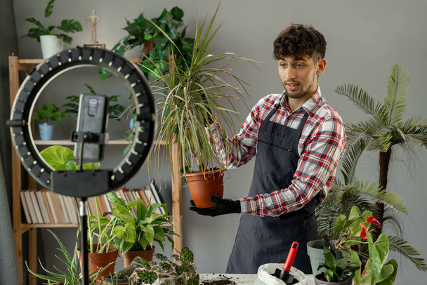 ソーシャルメディアのコンセプトに関するオンラインインフルエンサーの仕事. 若いブロガーの男はリビングルームに携帯電話が付いている陶器の植物が付いている家の庭のビデオを記録します. コピースペース - 写真・画像