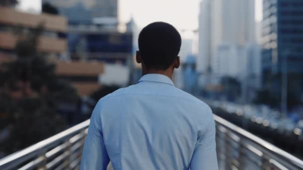 Back View of Young Successful African American Man in Shirt Walking Across Bridge in het zakendistrict. De rug van een zakenman die door de stad loopt op weg naar zijn werk. Het begrip mensen - Video