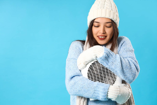 Mladá asijská žena v zimním oblečení s elektrickým ventilátorem topení na modrém pozadí - Fotografie, Obrázek