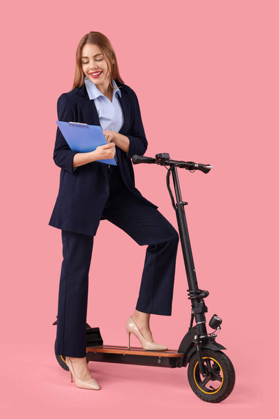 Portrait of young businesswoman with clipboard and kick scooter on pink background - Fotoğraf, Görsel