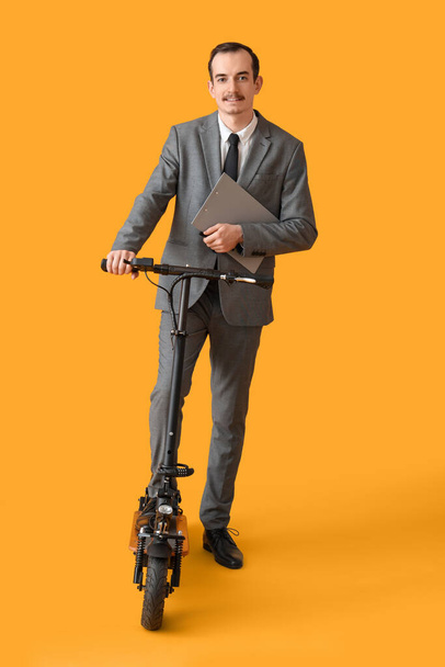 Handsome businessman with clipboard and kick scooter on yellow background - Фото, зображення