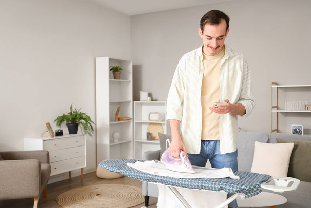 Young man with mobile phone ironing t-shirt on board at home - Fotoğraf, Görsel