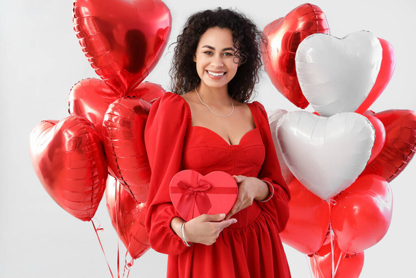 Young African-American woman with gift and balloons on light background. Valentine's Day celebration - Φωτογραφία, εικόνα