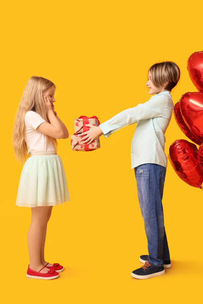 Little boy greeting girl with gift on yellow background. Valentine's Day celebration - Zdjęcie, obraz