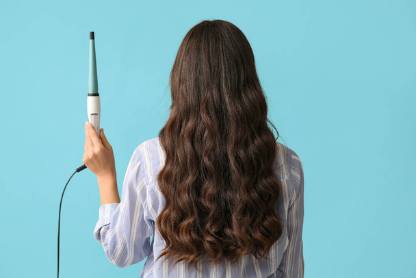 Beautiful young woman with curling iron on blue background, back view - Foto, Imagen