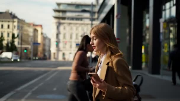 Молодая бизнесвумен в белой рубашке держит в руках мобильный телефон и читает сообщение при переходе возле современного здания в дневное время. Люди образ жизни  - Кадры, видео