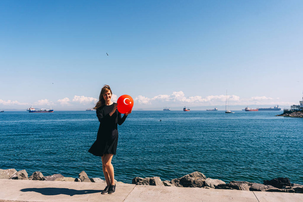 Ένα κορίτσι με ένα μαύρο φόρεμα με ένα κόκκινο μπαλόνι στο χέρι στην προβλήτα της Κωνσταντινούπολης. Ρωσίδα με ένα τούρκικο μπαλόνι στο βάθος της θάλασσας. μια γυναίκα μόνη στην Τουρκία - Φωτογραφία, εικόνα
