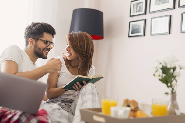 Belo jovem casal apaixonado se divertindo lendo e surfando na rede enquanto toma café da manhã na cama. Concentre-se na garota - Foto, Imagem