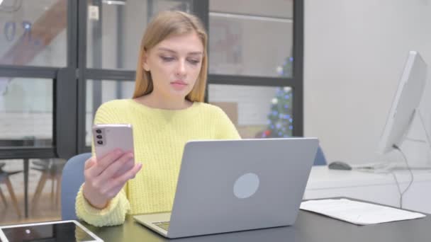 Νεαρή ξανθιά γυναίκα που εργάζονται σε φορητό υπολογιστή και τη χρήση Smartphone - Πλάνα, βίντεο