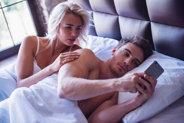 Krásný mladý pár leží v posteli, chlap používá chytrý telefon, dívka se dívá s výčitkou - Fotografie, Obrázek