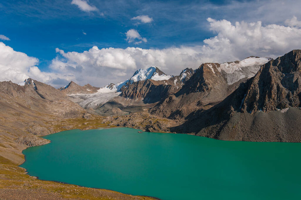 Alakel, a serene turquoise lake, is embraced by towering, rugged mountains with snow-capped peaks, under a vast and open blue sky, reflecting natures untouched splendor in the highlands - Photo, Image