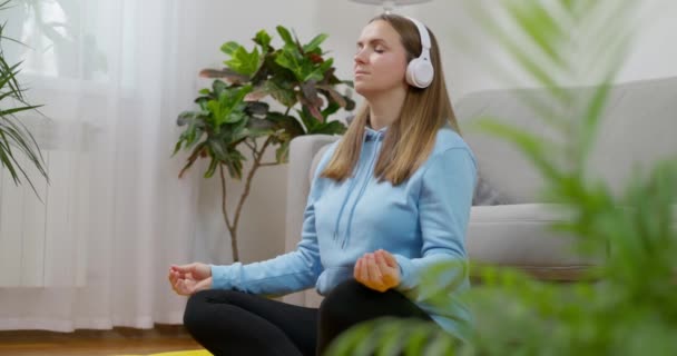 Nainen laittaa kuulokkeet täysin uppoutua meditaatioprosessiin istuessaan lootus aiheuttaa olohuoneen lattialle kotonaan. Laadukas 4k kuvamateriaalia - Materiaali, video
