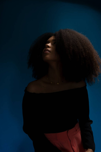 Half-closed portrait of young beautiful woman with black power hair looking up in low light. Isolated on blue background. - Foto, afbeelding