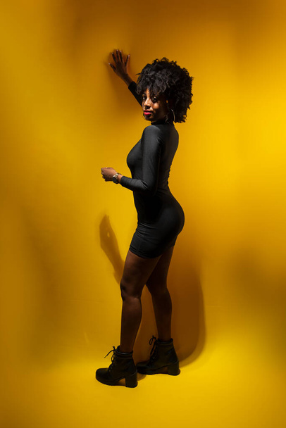 Fiatal érzéki nő fekete hatalmi haj, fekete ruhát visel támaszkodó sárga fal pózol egy fotó. - Fotó, kép