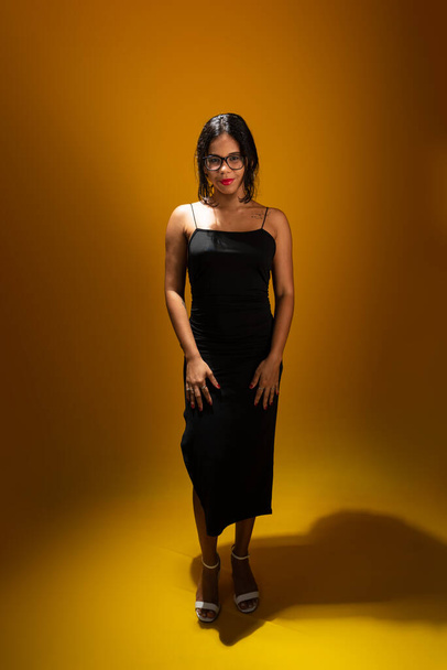 Retrato de bela sensual jovem morena vestindo roupas pretas usando óculos e posando para foto. Isolado sobre fundo amarelo. - Foto, Imagem