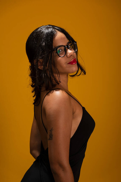 Retrato de bela sensual jovem morena vestindo roupas pretas usando óculos e posando para foto. Isolado sobre fundo amarelo. - Foto, Imagem