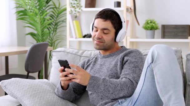 Junger hispanischer Mann wählt Musik in mobiler Anwendung. Person hört Musik über Kopfhörer, während sie auf dem Sofa sitzt. - Filmmaterial, Video