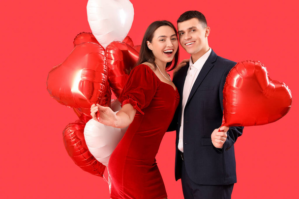 Νεαρό ζευγάρι με μπαλόνια σε σχήμα καρδιάς σε κόκκινο φόντο. Γιορτή του Αγίου Βαλεντίνου - Φωτογραφία, εικόνα