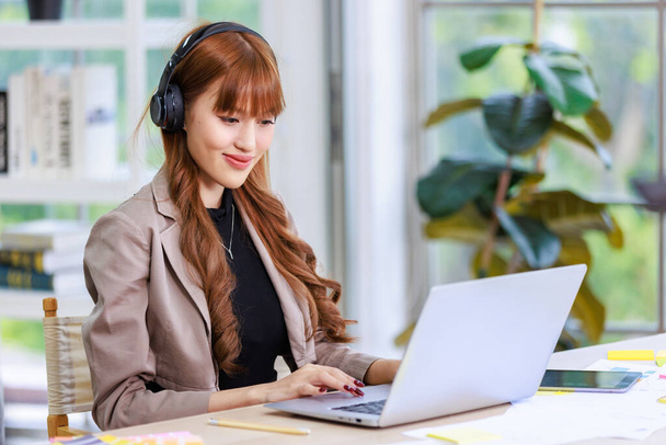 Asian professional successful young female businesswoman creative graphic designer in casual outfit sitting smiling wearing headphone listening to music playlist while working typing laptop computer. - Photo, Image