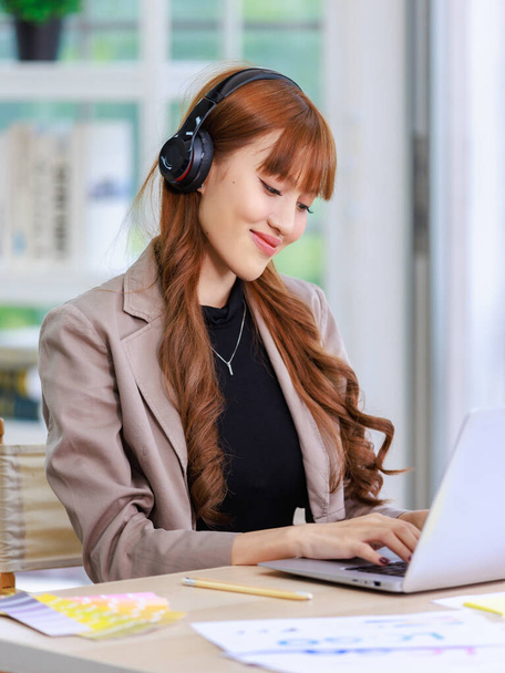 Azji profesjonalny sukces młoda kobieta bizneswoman kreatywny grafik w casual strój siedzi uśmiechnięty nosząc słuchawki słuchanie muzyki playlist podczas pracy pisania laptopa. - Zdjęcie, obraz