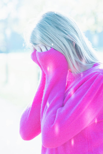 This artistic portrayal captures a young Caucasian woman in a moment of solitude, her face partially obscured by her raised fuchsia sweater, drawing the viewers focus to her hidden gaze. The high-key - Photo, Image