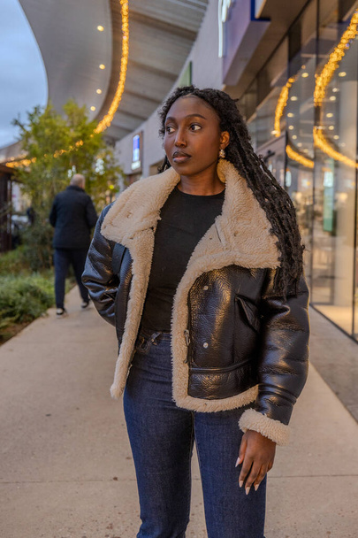Молода чорна жінка впевнено стоїть на міському тротуарі. Вона одягнена в повсякденне, стильне вбрання, з текстурованою чорною курткою для стрижки та темними джинсами, що пропонує прохолодний осінній день. Її волосся - Фото, зображення