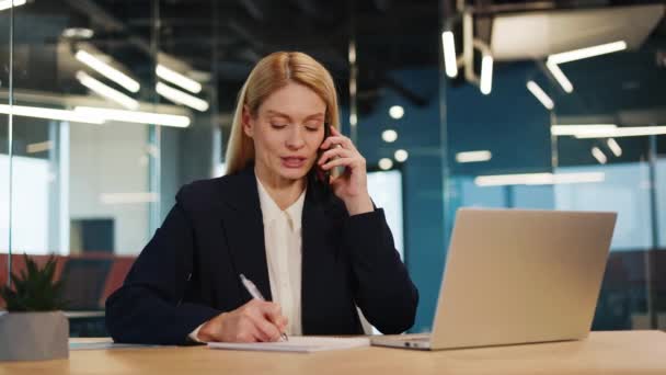 Όμορφη γυναίκα διευθύντρια γραφείου μιλάει στο τηλέφωνο κάθεται στον υπολογιστή στο γραφείο της. Όμορφη επιχειρηματίας μιλάει στο κινητό και εργάζεται με φορητό υπολογιστή freelancer έχοντας σημειώσεις κλήσης. - Πλάνα, βίντεο