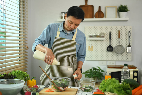 Šťastný muž středního věku v zástěře připravuje zdravý veganský salát v kuchyni. Zdravý životní styl koncept - Fotografie, Obrázek